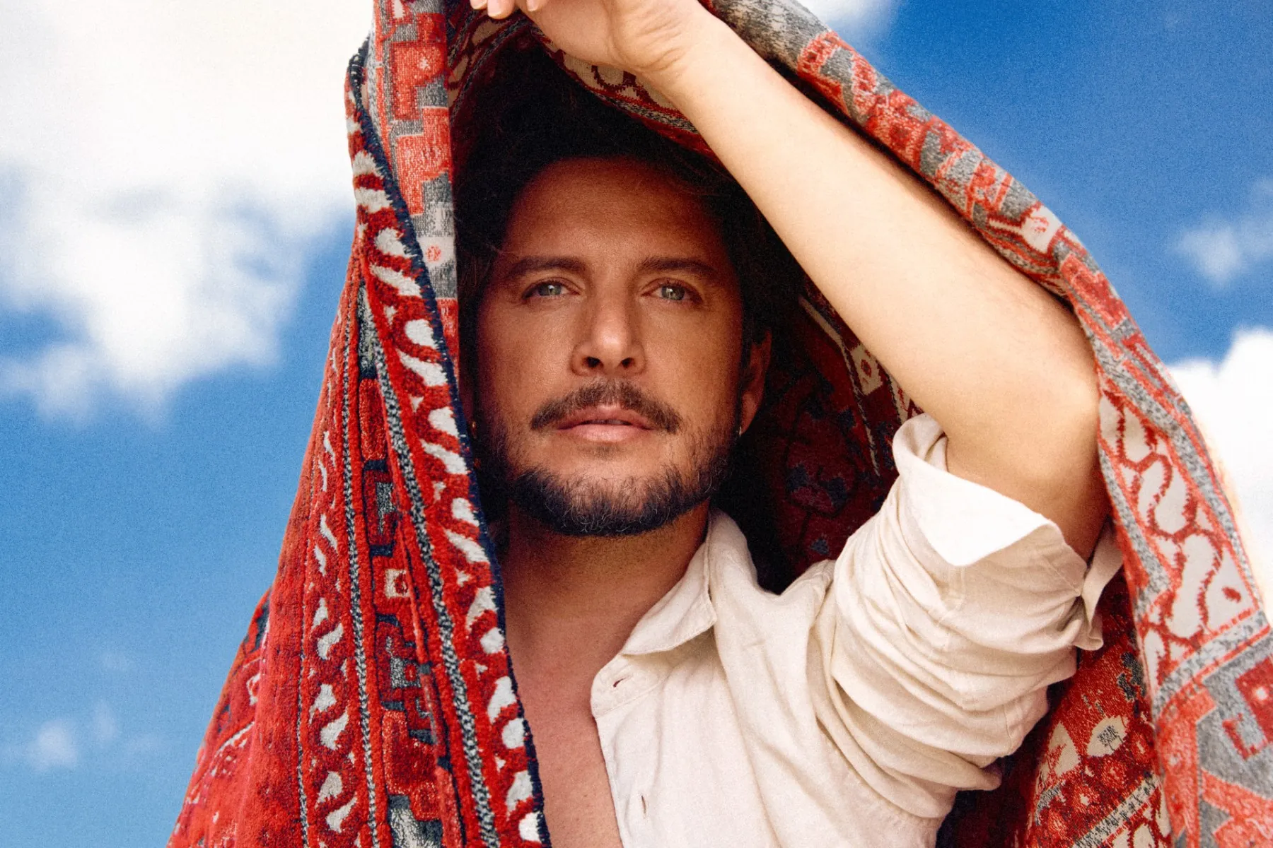 Manuel Carrasco con una alfombra kilim roja sobre la cabeza, contra un cielo azul cobalto.